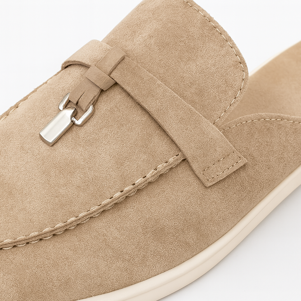 Suede mule loafer with a rounded toe, decorative strap, metal accent, and flat sole.