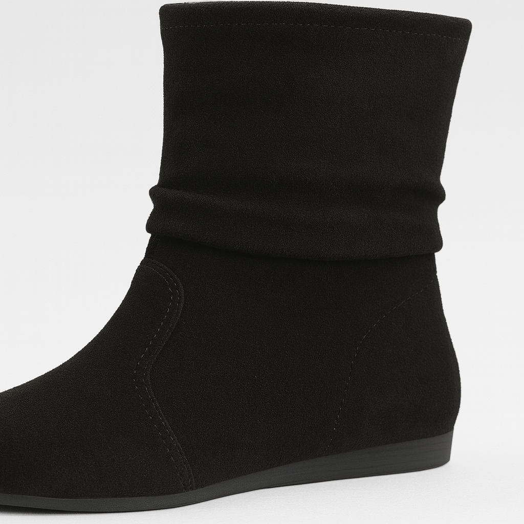 A pair of suede ankle boots with a slouchy, relaxed fit and flat soles. The boots feature a minimalist design with subtle stitching details and are shown worn on a model’s feet against a neutral background.