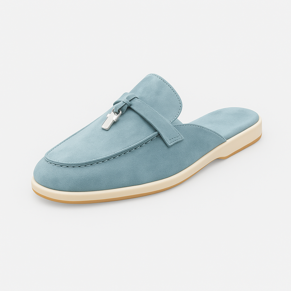Suede mule loafer with a rounded toe, decorative strap, metal accent, and flat sole.